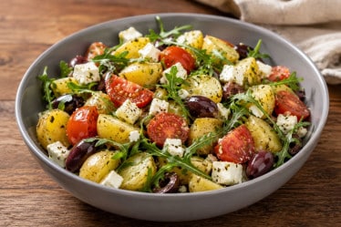 Mediterraner Kartoffelsalat ,it Tomate, Rucola, Feta und Zwiebeln in einer Schüssel auf einem Holztisch