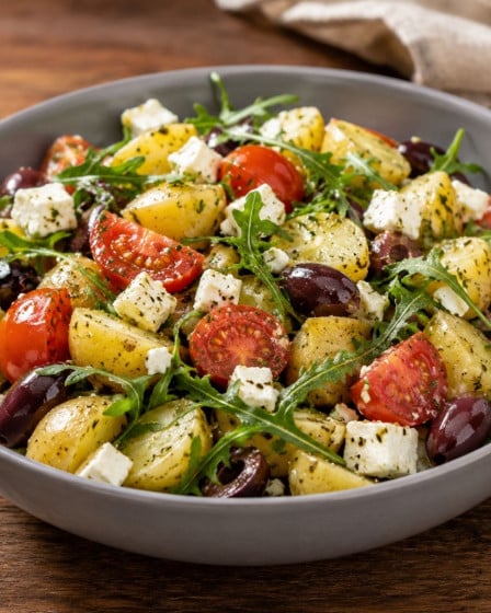 Mediterraner Kartoffelsalat ,it Tomate, Rucola, Feta und Zwiebeln in einer Schüssel auf einem Holztisch